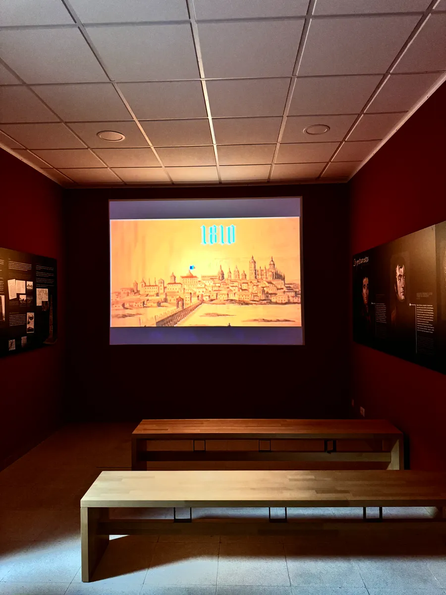 Sala audiovisual con bancos de madera frente a una proyección de una vista histórica de Salamanca.