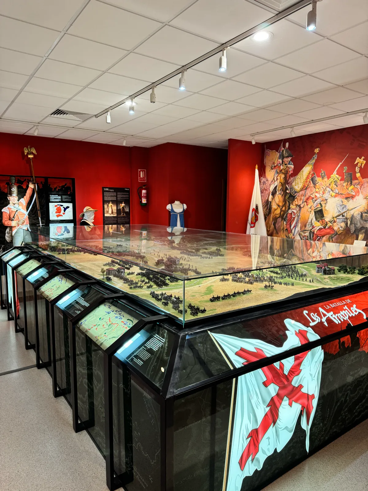 Gran maqueta de la batalla dentro de una vitrina alargada, con el terreno y miles de figuras desplegadas frente al mural del aula.