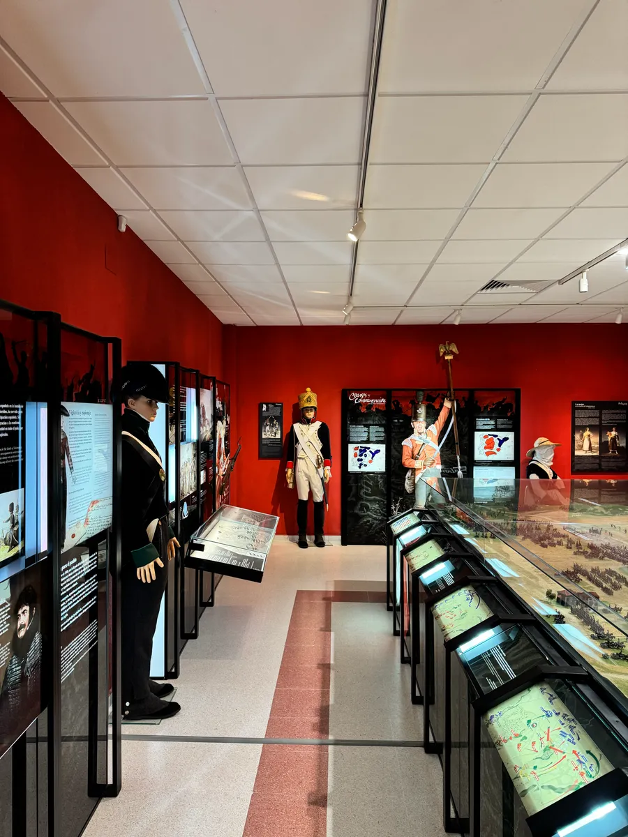 Sala del aula con paneles explicativos, figuras de soldados a tamaño real y vitrinas con maquetas, bajo paredes de color rojo.