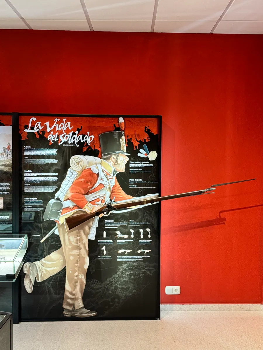 Panel «La vida del soldado» junto a una réplica de fusil con bayoneta, sobre la pared roja del aula.